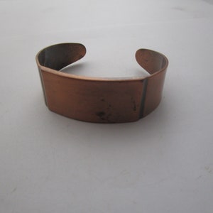 May include: A simple copper bracelet with a flat, rectangular design. The bracelet is open and has a slightly curved shape.