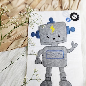 Könnte beinhalten: Ein grauer Roboter mit blauen Armen und einem gelben Blitz auf der Brust. Der Roboter hat einen blauen Bildschirm mit einer Wellenlinie. Der Roboter ist auf einen weißen Stoff gestickt.