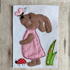 Könnte beinhalten: Ein braunes Häschen mit einem rosa gepunkteten Kleid und einem rosa Schmetterling auf dem Kopf. Das Häschen steht auf einem weißen Hintergrund mit einem grünen Grasfleck und einem roten Marienkäfer.