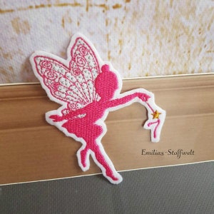 Könnte beinhalten: Rosa gestickte Fee mit weißen Flügeln, die einen goldenen Sternenzauberstab hält. Die Fee befindet sich auf einer Holzoberfläche mit dem Text "Emilias-Stoffwelt" darunter.