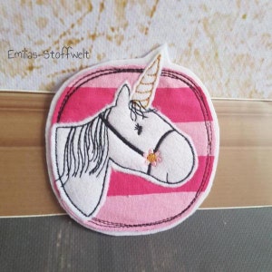 Könnte beinhalten: Ein gestickter Stoff-Patch mit einem weißen Einhorn mit goldenem Horn auf einem rosa-weiß gestreiften Stoff. Das Einhorn hat eine schwarze Mähne und einen schwarzen Schweif und eine rosa Blume auf der Nase.