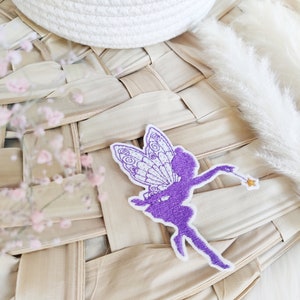 Könnte beinhalten: Lilafarbener gestickter Bügelaufkleber einer Fee mit Flügeln und einem Zauberstab. Die Fee ist vor einem weißen Hintergrund silhouettiert.