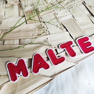 Könnte beinhalten: Rote, gestickte Aufnäher mit dem Namen "MALTE" mit weißem Rand, angeordnet auf einer gewebten, hellbraunen Oberfläche. Zarte weiße Blüten und grüne Stiele sind auf der gewebten Oberfläche platziert.