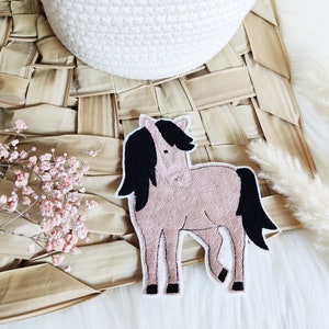 Könnte beinhalten: Ein rosa und schwarzer Filz-Pferde-Applik. Das Pferd steht auf einer geflochtenen Strohmatte.