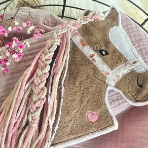 Könnte beinhalten: Ein brauner Plüschpferdekopf mit einer geflochtenen Mähne in Rosa und Weiß. Das Pferd trägt ein weißes Zaumzeug mit rosa floralen Akzenten und ein rosa Herz auf der Brust.