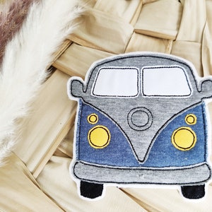 Könnte beinhalten: Eine graue und blaue Stoffapplikation eines Vintage-Vans mit gelben Scheinwerfern. Der Van befindet sich auf einem gewebten Hintergrund.