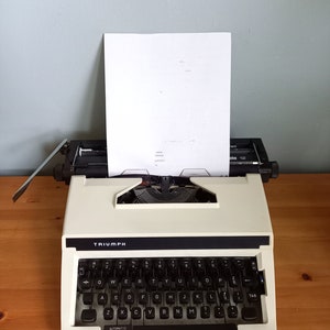 May include: A white Triumph typewriter with a black keyboard and a sheet of paper in the carriage. The typewriter has a black label with the word "TRIUMPH" on it. The keyboard has keys for letters, numbers, and symbols. The paper is blank.