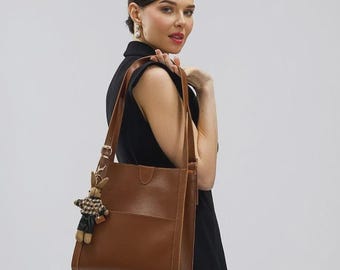 May include: A brown leather tote bag with a matching shoulder strap and a small decorative charm. The bag has a rectangular shape with a front pocket. The person is wearing a black sleeveless top and black knee-high boots.