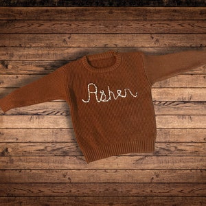 Coffee Brown colored Sweater with a natural white cursive embroidered name Asher  on wooden background