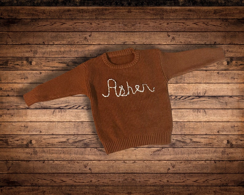 Coffee Brown colored Sweater with a natural white cursive embroidered name Asher  on wooden background