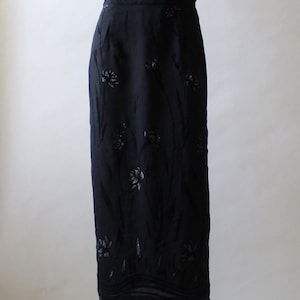 May include: A black maxi skirt with a floral pattern in silver. The skirt has a fitted silhouette and a slight flare at the hem.