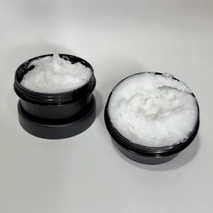 May include: Two black containers with white creamy substance inside. The containers are made of plastic and have a screw-on lid.