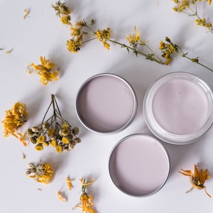 May include: Three round containers of skin care product with a light pink colour. The containers are made of metal and have a silver lid. The containers are on a white surface.