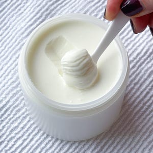 May include: A white plastic tub filled with a thick, creamy white substance. A white spoon scoops out a dollop of the cream, showing its texture. The tub is on a white, textured surface.