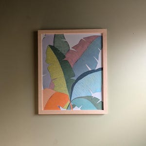 May include: A framed art print featuring a colorful illustration of tropical banana leaves. The leaves are depicted in shades of green, blue, pink, and orange, set against a light background. The artwork is encased in a light-colored wooden frame.