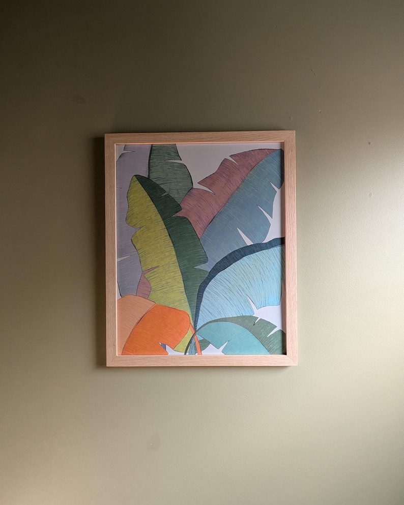 May include: A framed art print featuring a colorful illustration of tropical banana leaves. The leaves are depicted in shades of green, blue, pink, and orange, set against a light background. The artwork is encased in a light-colored wooden frame.