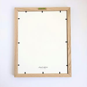May include: Back of a rectangular wooden picture frame with a white interior. The frame is made of light-colored wood and has black clips to hold the backing in place. The brand name "AnneCrafted" is printed in a script font on the lower right.