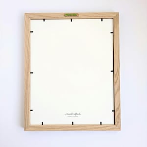 May include: A rectangular picture frame with a light brown wood grain finish, viewed from the back. The frame features a white backing board secured by black metal clips and a central metal hanging bracket. The text AnneCrafted is printed on the back.