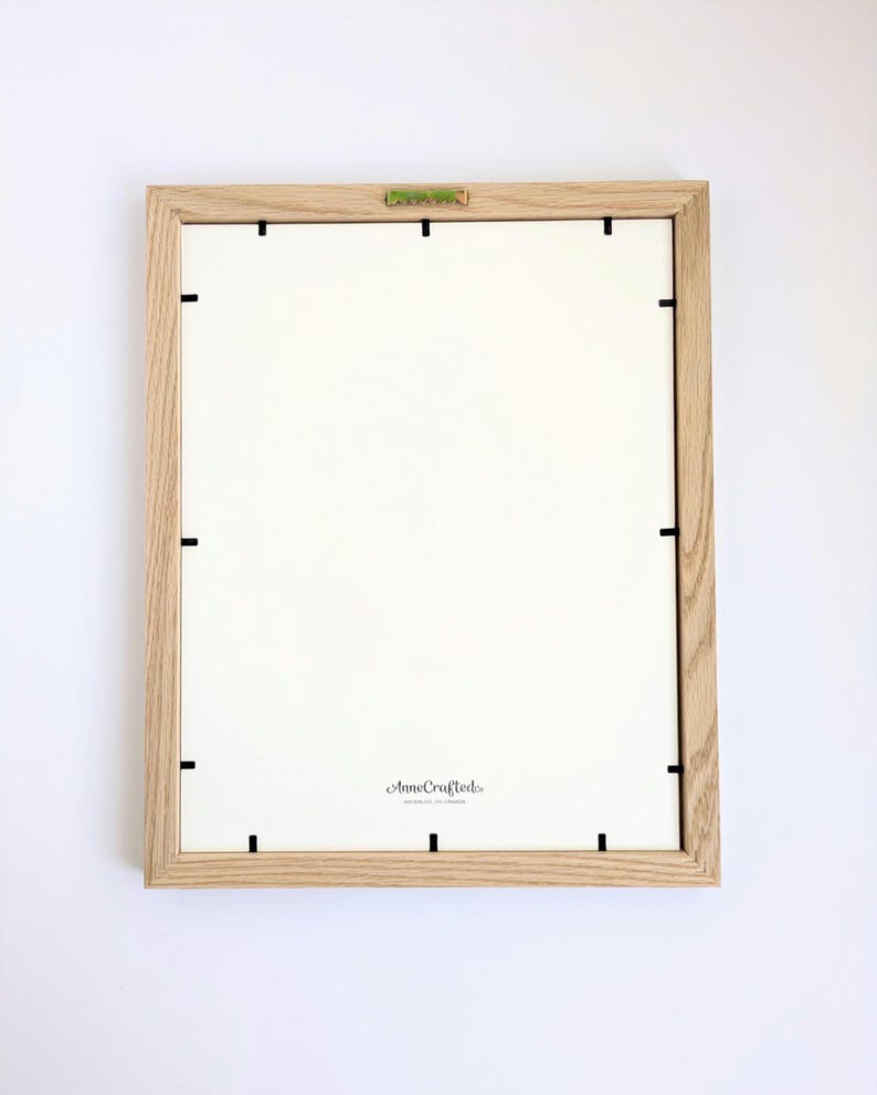 May include: A rectangular picture frame with a light brown wood grain finish, viewed from the back. The frame features a white backing board secured by black metal clips and a central metal hanging bracket. The text AnneCrafted is printed on the back.
