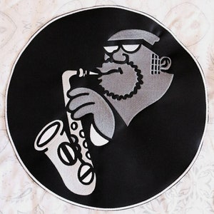 May include: Black and white embroidered patch of a cartoon man playing a saxophone. The man is wearing a hat and sunglasses.