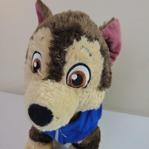 May include: Brown and tan plush dog toy wearing a blue jacket. The dog has large, expressive eyes and floppy ears.  The plush toy is soft and cuddly, suitable for children.  It's a great addition to any child's toy collection. The dog's fur is a mix of light tan and dark brown, giving it a realistic look. The blue jacket adds a pop of color to the toy.