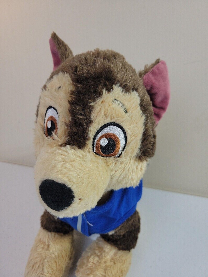 May include: Brown and tan plush dog toy wearing a blue jacket. The dog has large, expressive eyes and floppy ears.  The plush toy is soft and cuddly, suitable for children.  It's a great addition to any child's toy collection. The dog's fur is a mix of light tan and dark brown, giving it a realistic look. The blue jacket adds a pop of color to the toy.