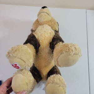 May include: Tan and brown plush Beanie Boo's dog toy. The dog has a stitched brown nose and black eyes.  The toy's paws have a small, round, red paw print logo. The plush toy is shown from above, lying on its back with its legs in the air.