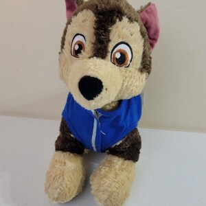 May include: Plush toy dog wearing a blue jacket. The dog is light brown and tan with dark brown fur on its back and head. It has large, expressive eyes and a friendly face. The plush toy is sitting on a white surface.  The toy is soft and cuddly, suitable for children. This is a great gift for fans of the Paw Patrol cartoon.