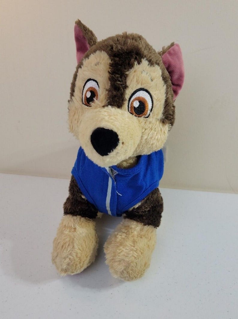 May include: Plush toy dog wearing a blue jacket. The dog is light brown and tan with dark brown fur on its back and head. It has large, expressive eyes and a friendly face. The plush toy is sitting on a white surface.  The toy is soft and cuddly, suitable for children. This is a great gift for fans of the Paw Patrol cartoon.