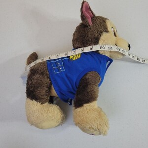 May include: Brown and tan plush Paw Patrol Ryder's pup, Chase, wearing a blue vest with a paw print and badge. The plush toy is approximately 12 inches long and is shown from the side.  The plush toy's fur is soft and fluffy. The vest has a velcro closure.  The toy is suitable for children.