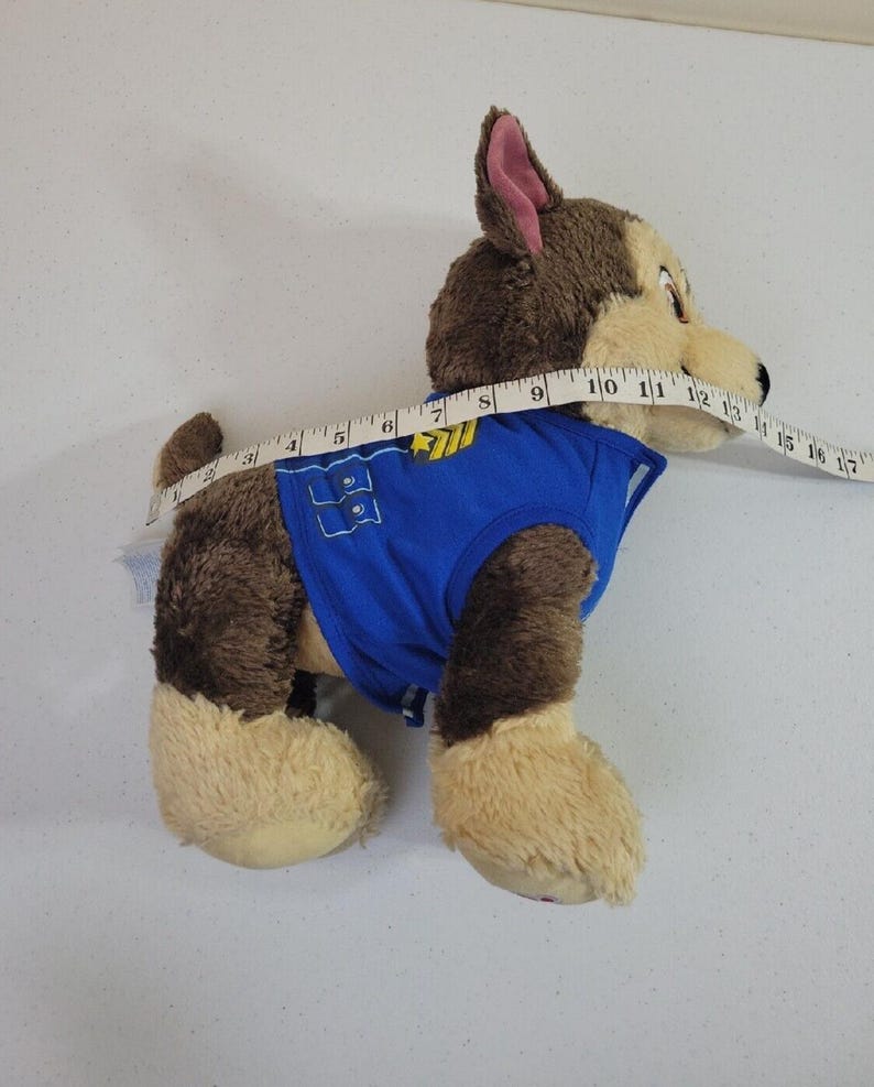 May include: Brown and tan plush Paw Patrol Ryder's pup, Chase, wearing a blue vest with a paw print and badge. The plush toy is approximately 12 inches long and is shown from the side.  The plush toy's fur is soft and fluffy. The vest has a velcro closure.  The toy is suitable for children.