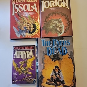 May include: Four fantasy novels by Steven Brust are displayed. The covers feature illustrations of a dragon, a wolf, and a man. The titles include "Issola", "Jorich", "Athyra", and "The Paths of the Dead". The books are in various sizes and colors.
