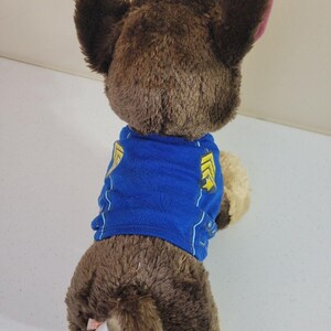 May include: Brown plush Paw Patrol Chase toy wearing a blue shirt with yellow accents. The toy is shown from the back, showcasing its soft fur and detailed features.  A tag is visible on the toy's rear, indicating the manufacturer. The plush toy is a collectible item for fans of the popular children's show.