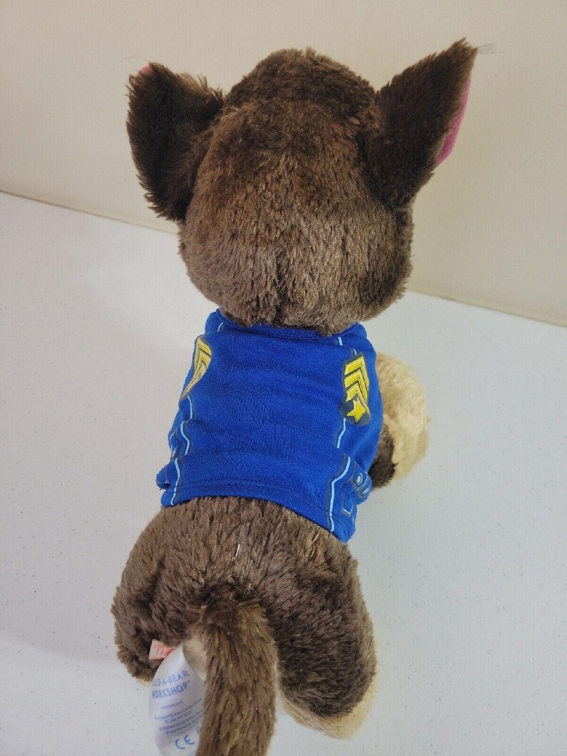 May include: Brown plush Paw Patrol Chase toy wearing a blue shirt with yellow accents. The toy is shown from the back, showcasing its soft fur and detailed features.  A tag is visible on the toy's rear, indicating the manufacturer. The plush toy is a collectible item for fans of the popular children's show.