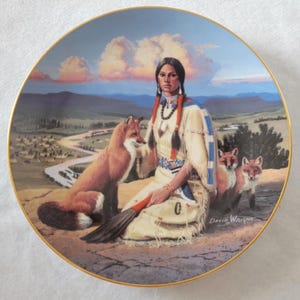 Puede incluir: Plato decorativo con borde dorado, que representa a una mujer nativa americana con vestimenta tradicional y zorros. La escena incluye un río, un pueblo y un atardecer. Firmado "David Wright".