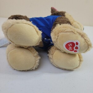 May include: Brown and tan plush dog toy wearing a blue shirt. The toy has a paw print logo on its bottom.  The dog is lying on its side. The logo says 'BAB'.