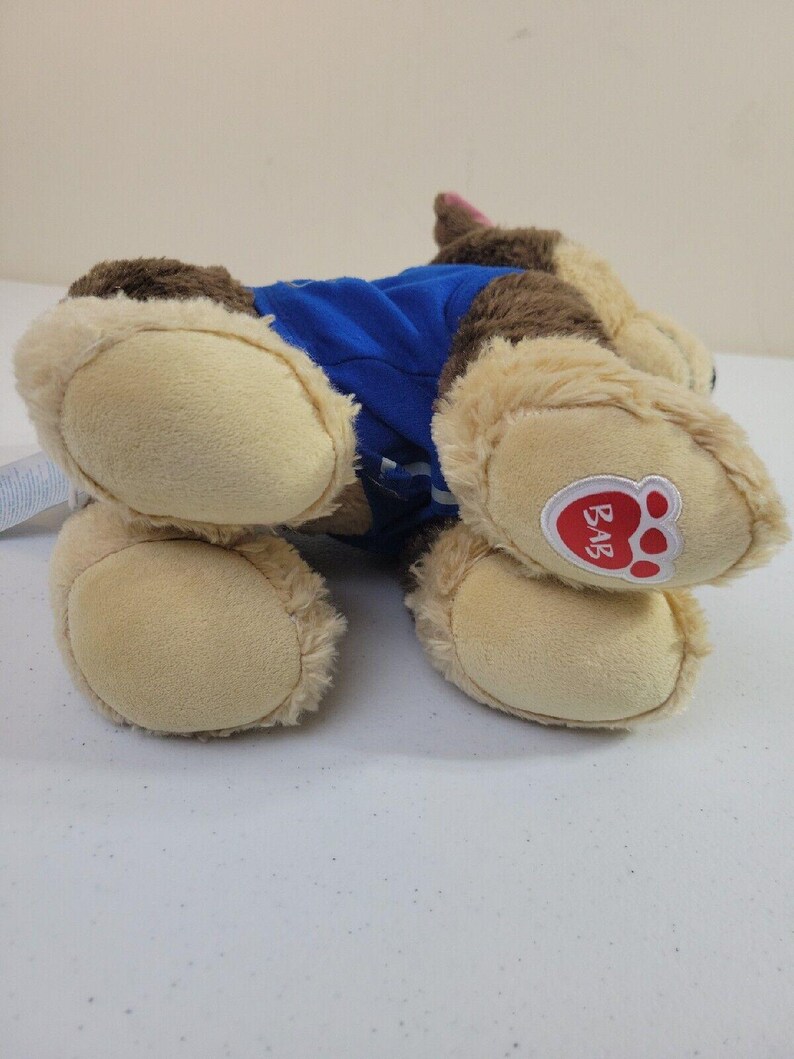 May include: Brown and tan plush dog toy wearing a blue shirt. The toy has a paw print logo on its bottom.  The dog is lying on its side. The logo says 'BAB'.
