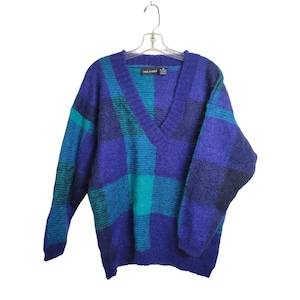 May include: A vintage Paul Harris V-neck sweater in a plaid pattern. The sweater features shades of blue and teal. The sweater is hanging on a clear plastic hanger. The sweater is made of a soft, fuzzy material.