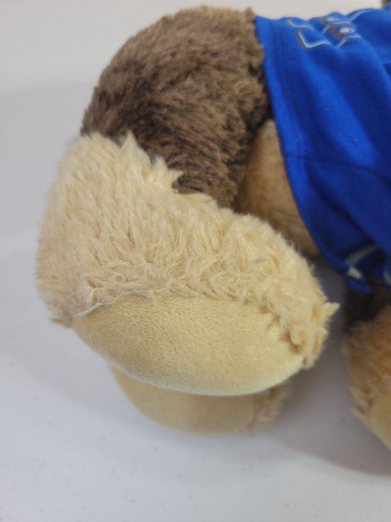 May include: Brown and tan plush dog toy wearing a blue shirt. The dog is lying down, and its paws are visible. The fur is soft and fluffy. The shirt has some text on it that is not clearly visible. The toy is suitable for children and is made of high-quality materials.