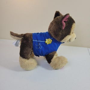 May include: Side view of a brown and tan plush toy dog wearing a blue uniform. The uniform has yellow accents and features a badge. The dog is a Paw Patrol character, possibly Chase. The toy is soft and cuddly, suitable for children.  The plush toy is shown standing on a light-colored surface.