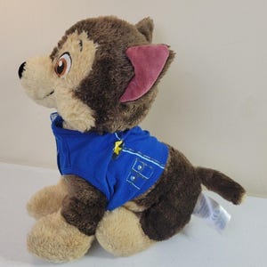 May include: Brown and tan plush Paw Patrol Ryder's pup, Chase, wearing a blue police uniform. The plush toy is sitting and facing to the left.  The plush toy has brown fur, tan paws and a pink inner ear. The blue uniform has a yellow star emblem.