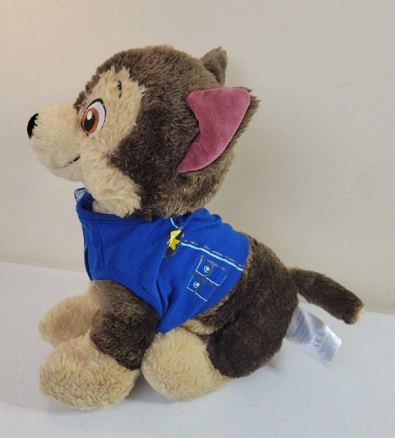 May include: Brown and tan plush Paw Patrol Ryder's pup, Chase, wearing a blue police uniform. The plush toy is sitting and facing to the left.  The plush toy has brown fur, tan paws and a pink inner ear. The blue uniform has a yellow star emblem.