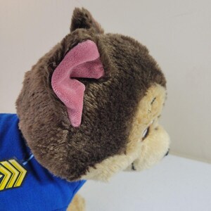 May include: Brown and tan plush dog toy wearing a blue shirt with yellow chevron detail. The dog has pink inner ears and is in profile view.  The toy is a Paw Patrol character.