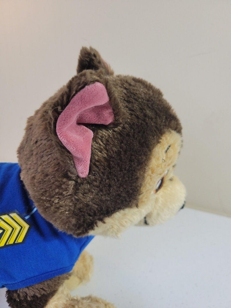 May include: Brown and tan plush dog toy wearing a blue shirt with yellow chevron detail. The dog has pink inner ears and is in profile view.  The toy is a Paw Patrol character.