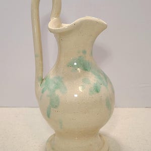 May include: A small, off-white ceramic pitcher with a curved handle and a flared spout. The pitcher is decorated with light green leaf-like patterns. It has a round base and a height of approximately 6 inches.