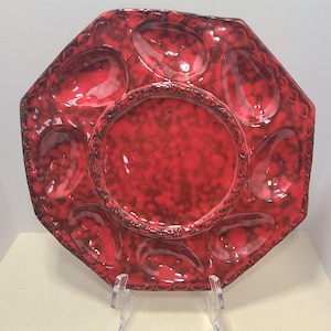May include: A red, octagonal ceramic oyster plate with six oval indentations and a central circular well. The plate has a textured surface and a decorative border. The plate is displayed on a clear acrylic stand.