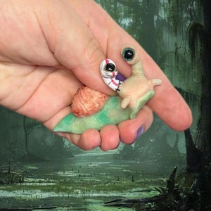 May include: A small, sculpted snail figurine with a pale green body and a textured, brain-like pink shell. The snail features two prominent, stalked eyes with black pupils, one of which is wrapped in a white bandage with red stitching details.