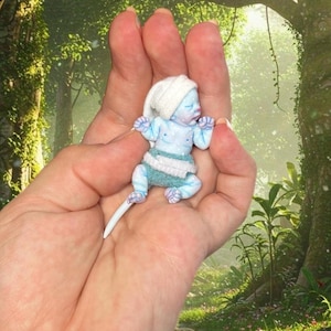 May include: A miniature figurine of a pale blue creature with white spots, wearing a white cap and a teal diaper. The creature has a long, thin white tail extending from its lower back and is depicted in a sleeping position with closed eyes.