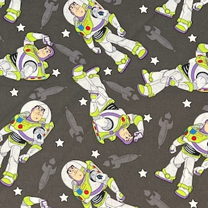 May include: A gray fabric with a repeating pattern of a cartoon space ranger. The space ranger is wearing a white and green spacesuit with a yellow helmet and a red star on his chest. The background is a dark gray with white stars.