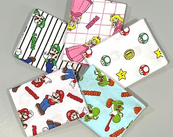 May include: Five fabric squares with different designs featuring characters from the video game Super Mario Bros. The squares are white with colorful designs. One square features a pink princess with a crown, another square features a green character with a mustache, and another square features a red character with a mustache. The squares are all different sizes and shapes.